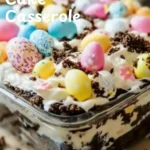 Eazy Fun Easter Dirt Cake Casserole % A glass dish filled with Dirt Cake Casserole, decorated with pastel chocolate eggs and cookie crumbles, with colorful spoons in the background.